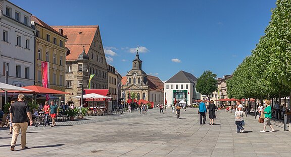 Ein belebter Platz in einer Stadt mit historischen Gebäuden. Cafés mit roten Sonnenschirmen laden zum Verweilen ein. Menschen spazieren oder sitzen, während Bäume entlang des Platzes Schatten spenden. Der Himmel ist klar und blau.