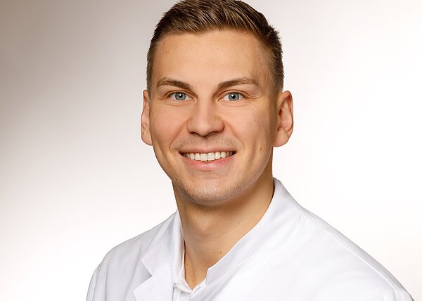 A smiling young man with short hair is wearing a white lab coat. He has blue eyes and appears friendly and approachable. The background is a soft, neutral gradient.