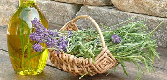 Glasflasche mit goldenem Öl neben kleinem Weidenkorb, gefüllt mit frischen Lavendelzweigen und violetten Blüten.