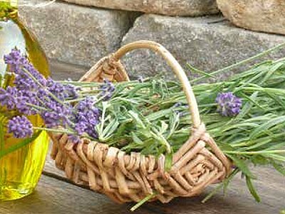Glasflasche mit goldenem Öl neben kleinem Weidenkorb, gefüllt mit frischen Lavendelzweigen und violetten Blüten.