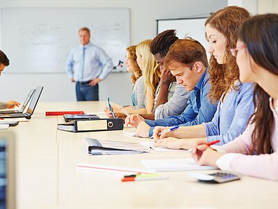Eine Gruppe von Studierenden sitzt an einem Tisch in einem Klassenraum. Sie konzentrieren sich auf ihre Laptops und Notizen, während ein Lehrer im Hintergrund steht und die Klasse leitet. Einige Schüler schreiben mit Stiften, andere sehen auf ihre Bildschirme.