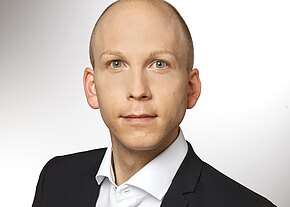 Ein junger Mann mit kurzem, glattem Kopf trägt einen schwarzen Anzug und ein weißes Hemd. Er schaut direkt in die Kamera mit neutrale Miene. Der Hintergrund ist hell und unscharf, wodurch der Fokus auf seinem Gesicht liegt.