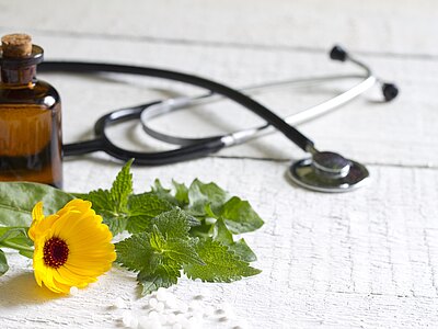 Braunes Medizinfläschchen, Stethoskop, Ringelblume und Kräuter neben weißen Globuli auf hellem Holztisch – Symbol für Naturheilkunde bzw. Verbindung von Schulmedizin und pflanzlicher/alternativer Behandlung.