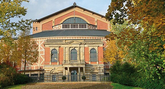 Frontansicht eines großen Opern- und Theatergebäudes aus rotem Backstein mit klassizistischer Sandsteinfassade, Rundbogenfenstern, zentralem Eingang und Giebel. Das Gebäude ist von herbstlich gefärbten Bäumen eingerahmt.