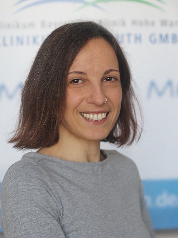 Porträtfoto: Eine lächelnde Frau mit kinnlangem braunem Haar und Nasenring blickt in die Kamera. Sie trägt ein graues Oberteil. Der Hintergrund ist unscharf und neutral.