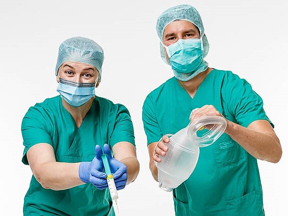 Zwei medizinische Fachkräfte in grünen Scrubs und OP-Hauben stehen zusammen. Die Frau hält eine Spritze und zeigt darauf, während der Mann einen medizinischen Behälter in der Hand hält. Beide tragen Mund-Nasen-Schutz und schauen freundlich in die Kamera.