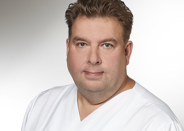 Porträt eines erwachsenen Mannes in weißer medizinischer Kleidung vor neutralem Hintergrund. Er blickt ruhig in die Kamera; Darstellung eines Gesundheitsmitarbeiters.