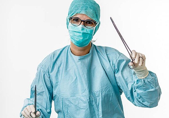 Medizinische Fachkraft in OP-Kleidung vor hellem Hintergrund. Die Person trägt Haube, Maske, Handschuhe und Brille und hält eine chirurgische Schere sowie eine Klemme erhoben, bereit für einen Eingriff.
