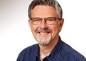 Christian Pfeiffer Ein lächelnder Mann mit grauem Haar und Brille steht vor einem hellen Hintergrund. Er trägt ein dunkelblaues Hemd und hat einen kurzen, gepflegten Bart. Der Ausdruck auf seinem Gesicht vermittelt Freundlichkeit und Offenheit.