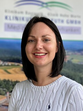 Porträt einer lächelnden Frau mit schulterlangem, dunklem Haar, in weißem Strickpullover. Sie blickt direkt in die Kamera vor unscharfem, nebensächlichem Hintergrund. Schwerpunkt: freundliches Porträtfoto.