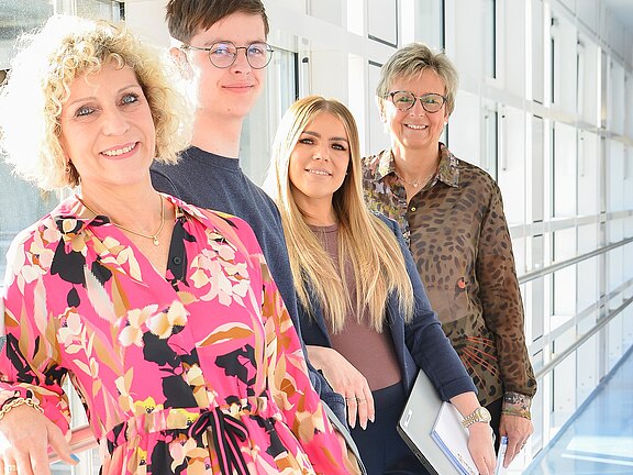 Vier Personen unterschiedlichen Alters stehen nebeneinander in einem hellen Flur und lächeln in die Kamera. Vorn eine Frau im bunten Kleid, dahinter ein junger Mann, eine Frau mit langen Haaren und eine ältere Frau mit Unterlagen. Eindruck: freundliches, professionelles Team.