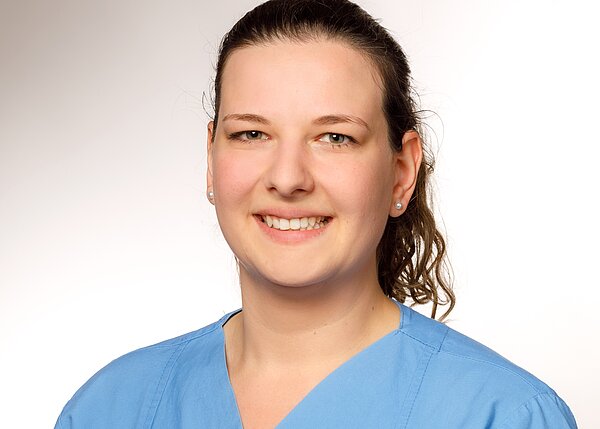 A smiling woman wearing a blue medical scrub shirt is depicted. She has long, curly hair pulled back and is wearing small earrings. The background is lightly shaded, emphasizing her friendly expression.