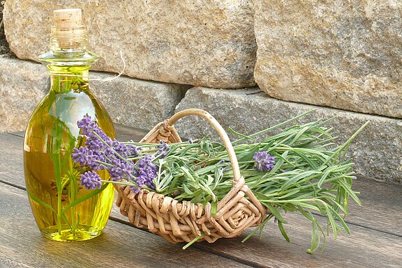 Glasflasche mit goldenem Öl neben einem kleinen geflochtenen Korb voller frisch geschnittener Lavendelzweige. Violette Blüten auf langen grünen Stielen. Eindruck von Natur, Kräuterküche oder Aromatherapie.