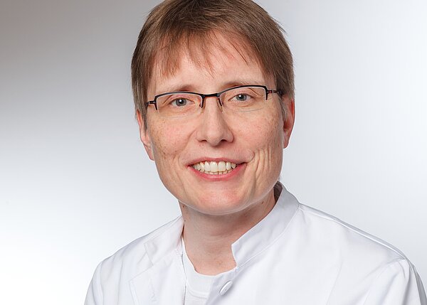 A smiling person with short hair and glasses is wearing a white medical coat. The background is light and neutral, emphasizing the subject’s friendly expression.