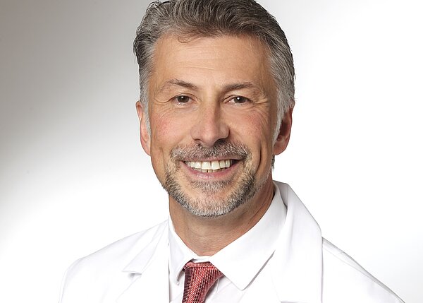 A smiling male doctor with short gray hair and a neatly trimmed beard wears a white lab coat over a dress shirt and tie. He exudes a professional demeanor, standing against a light background.