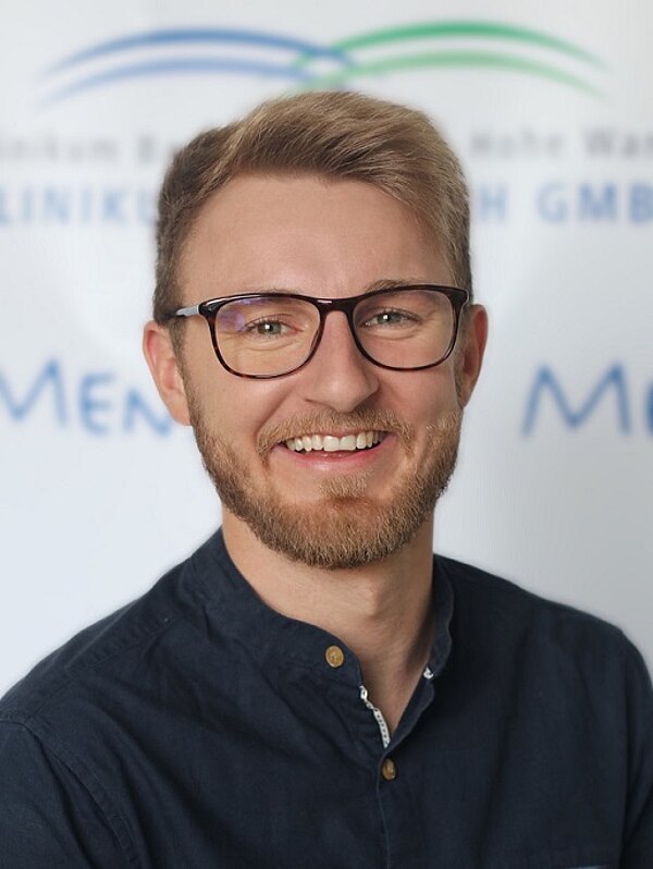 Porträtfoto eines lächelnden jungen Mannes mit Brille und kurzem Bart. Er trägt ein dunkelblaues Hemd und blickt direkt in die Kamera vor neutralem, unscharfem Hintergrund.