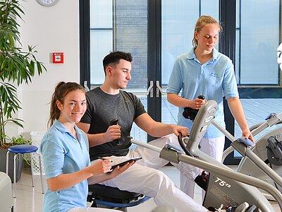 In einem modernen Trainingsraum helfen zwei Physiotherapeuten einem Mann, der an einem Trainingsgerät sitzt. Eine der Therapeutinnen notiert etwas. Alle tragen sportliche Kleidung und wirken freundlich, während sie sich auf das Training konzentrieren.