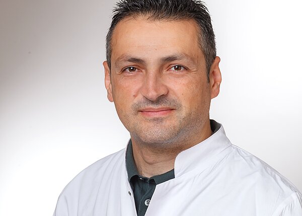 A male medical professional with short black hair smiles at the camera. He is wearing a white lab coat over a dark shirt. The background is a light gradient, enhancing his professional appearance.