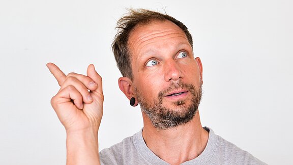 Ein Mann vor hellem Hintergrund blickt nach oben links und zeigt mit erhobenem Zeigefinger dorthin, als wolle er auf etwas hinweisen oder eine Idee haben.
