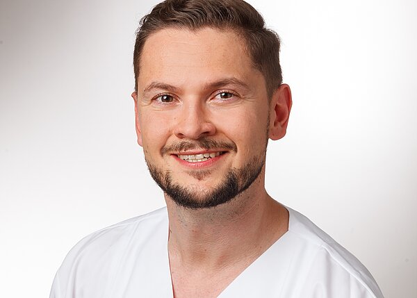 A smiling male healthcare professional is wearing a white scrub top. He has short hair and a well-groomed beard. The background is bright and plain, emphasizing the subject's friendly expression.