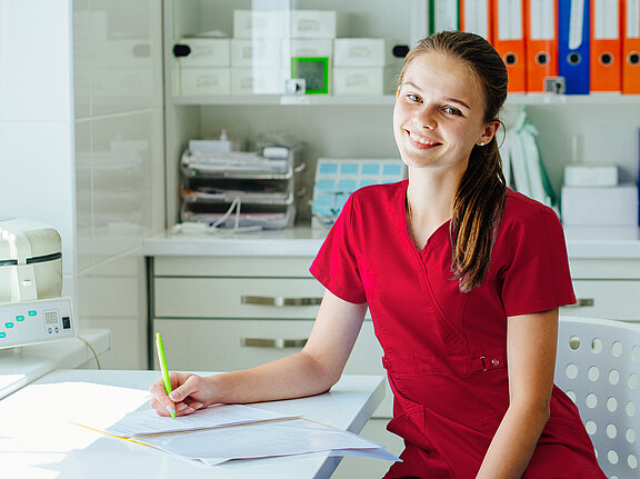 Junge medizinische Fachkraft in roter Arbeitskleidung sitzt lächelnd an einem Schreibtisch in einer Praxis und füllt Unterlagen aus.