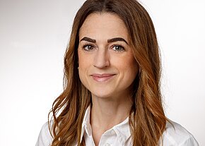 Nahaufnahme einer lächelnden Frau mit langen, lockigen braunen Haaren. Sie trägt ein weißes Hemd und hat eine freundliche Ausstrahlung. Der Hintergrund ist hell und neutral.