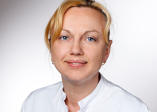 A woman with light hair and blue eyes smiles gently. She is wearing a white medical uniform. The background is softly blurred, emphasizing her face and expression, suggesting a professional healthcare setting.