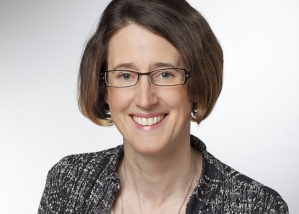 Portrait of a smiling adult with short brown hair and glasses, wearing a patterned blazer and necklace, against a plain light background.