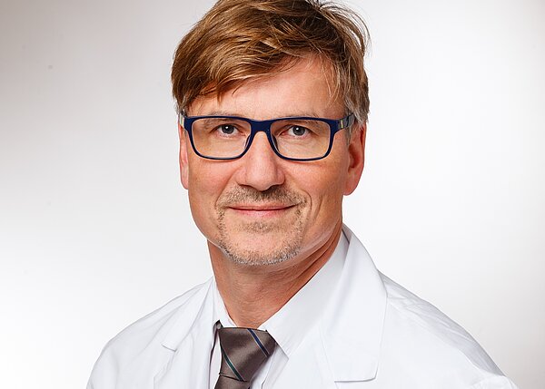 A man with short, light brown hair and glasses is wearing a white lab coat over a collared shirt and tie. He is smiling gently, conveying a professional and approachable demeanor, against a plain, light background.