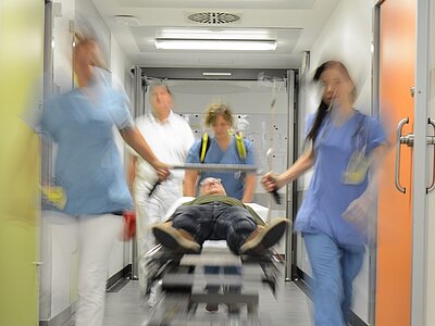 Im Krankenhausflur schiebt ein Notfallteam einen Patienten auf einer Trage eilig Richtung Behandlungsraum. Die Bewegungsunschärfe betont die Dringlichkeit; medizinisches Personal in blauer Kleidung begleitet den Patienten mit Infusionsständer.