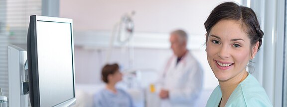 Lächelnde Pflegekraft im Vordergrund eines Krankenhauszimmers. Im Hintergrund spricht ein Arzt mit einer Patientin am Bett. Ein Monitor steht daneben. Szene vermittelt freundliche, professionelle Betreuung.