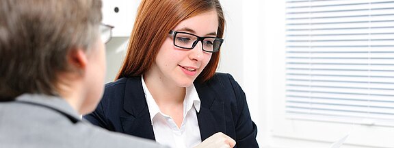 Two professionals in an office meeting. A woman with red hair and glasses reviews a document and explains it to a colleague across the desk.