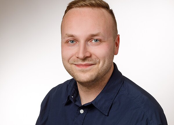 Porträtfoto eines lächelnden jungen Mannes mit kurzem blondem Haar. Er trägt ein dunkelblaues Hemd und blickt direkt in die Kamera. Heller, neutraler Hintergrund, Kopf-und-Schultern-Ausschnitt.