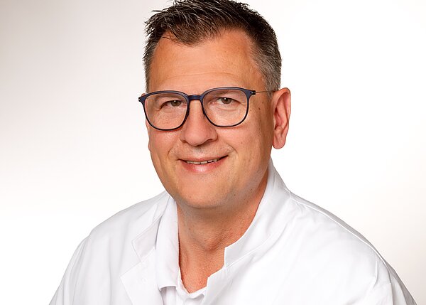 A smiling male doctor with short dark hair and glasses is shown. He is wearing a white lab coat and has a friendly expression. The background is light, emphasizing his face.
