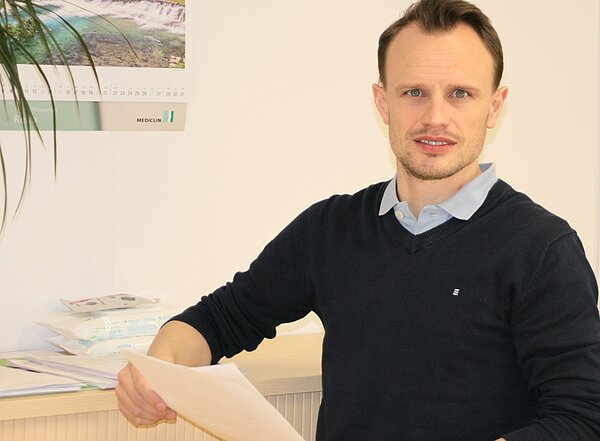 A man with short hair sits comfortably at a desk, holding a stack of papers. He wears a dark sweater over a light blue collared shirt. The background features a plant and a calendar displaying a scenic landscape.