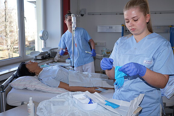 In einem Krankenhauszimmer liegt eine Patientin im Bett; zwei Pflegekräfte in hellblauer Kleidung bereiten eine Behandlung vor. Eine Person zieht blaue Handschuhe an, die andere arbeitet am Infusionsständer.