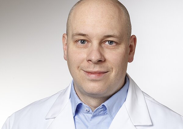 A close-up portrait of a man wearing a white lab coat over a blue shirt. He has a shaved head, blue eyes, and a slight smile, conveying a professional and friendly demeanor.