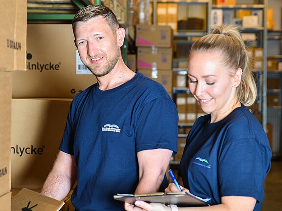 Zwei Personen in blauen T‑Shirts arbeiten im Lager zwischen Kartons. Eine Person hält einen Karton, die andere schreibt auf einem Klemmbrett; die Szene zeigt Inventur oder Kommissionierung.