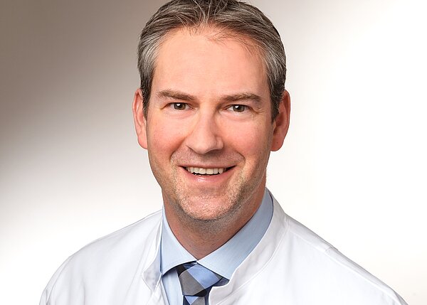 A smiling male doctor is wearing a white coat over a light blue dress shirt and patterned tie. He has short, neatly styled hair and is looking directly at the camera, conveying a friendly and approachable demeanor. The background is a soft, neutral tone.