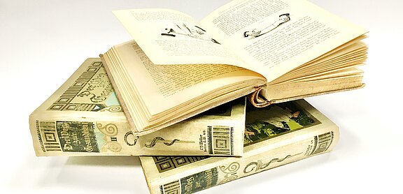 Drei alte, cremefarbene Bücher liegen gestapelt; das oberste ist aufgeschlagen und zeigt Text und Illustrationen. Dekorative Einbände im Retro-Stil, neutraler Hintergrund.