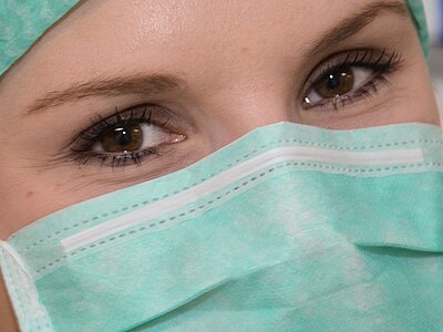 Nahaufnahme einer medizinischen Fachkraft mit grüner OP-Mütze und chirurgischer Maske; nur die Augen sind sichtbar. Aufmerksamer Blick, Symbol für Hygiene und Gesundheitsschutz.