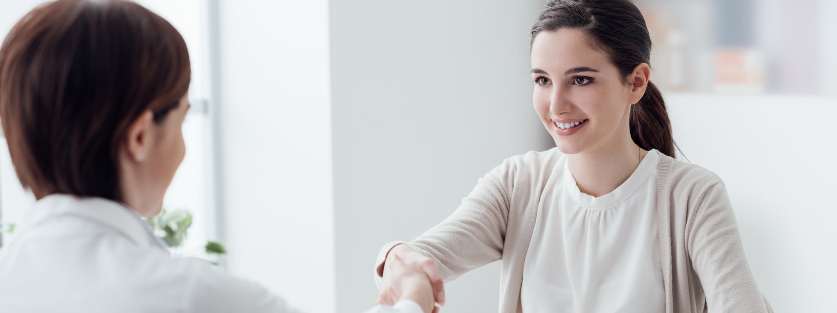 Zwei Frauen in einem hellen Raum schütteln sich die Hände. Eine Frau lächelt freundlich, während die andere ihr den Rücken zukehrt. Diese Geste deutet auf einen positiven Austausch oder ein Kennenlernen hin.