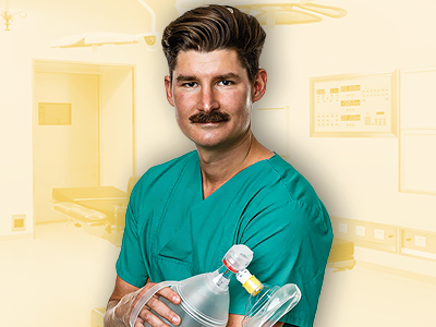 Ein junger Mann mit einem Schnurrbart trägt grüne OP-Kleidung und hält ein Beatmungsgerät in den Händen. Im Hintergrund sind verschwommene Elemente eines Krankenhausraums erkennbar, was die medizinische Umgebung unterstreicht.