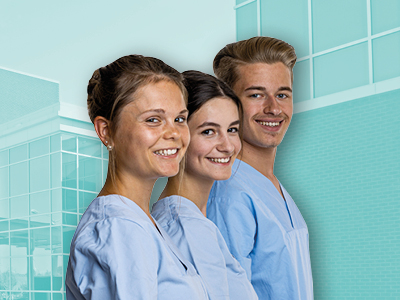 Drei junge Menschen in hellblauen Pflegekitteln stehen nebeneinander und lächeln vor einer modernen Krankenhausfassade.