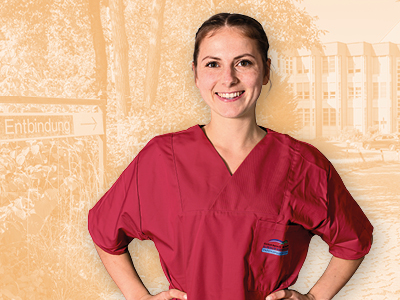 Lächelnde Frau in bordeauxroter Krankenhauskleidung steht mit Händen in den Hüften. Im Hintergrund ein Krankenhaus mit Schild „Entbindung“, Hinweis auf Geburtshilfe.
