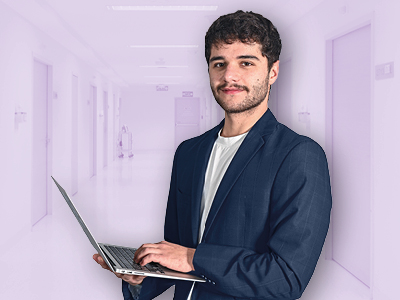 Fokus auf einen jungen Mann im dunkelblauen Blazer, der lächelnd an einem offenen Laptop arbeitet; heller Flur im Hintergrund.