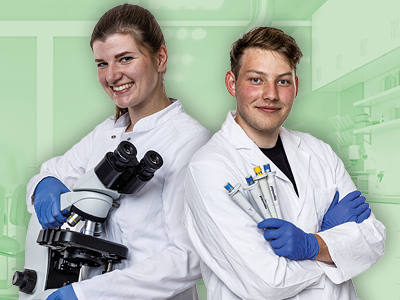 Zwei junge Labormitarbeitende in weißen Kitteln und blauen Handschuhen stehen nebeneinander. Die Person links hält ein Mikroskop und lächelt, die Person rechts hält mehrere Pipetten. Hintergrund: Laborumgebung. Motiv vermittelt professionelle Laborarbeit und Teamwork.