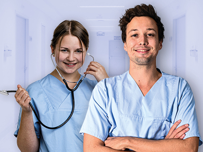 Zwei medizinische Fachkräfte in hellblauer OP-Kleidung stehen in einem Krankenhausflur. Die Frau lächelt und hält ein Stethoskop, der Mann steht daneben mit verschränkten Armen und lächelt. Das Bild vermittelt professionelle, freundliche Pflege.