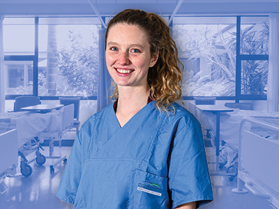 Porträt einer lächelnden Pflegekraft in blauer Dienstkleidung, stehend in einer Krankenhausstation mit Betten und Fenstern.