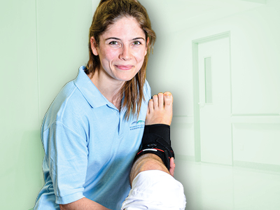 Physiotherapeutin im Klinikflur versorgt einen Patientenfuß: Sie befestigt eine schwarze Sprunggelenkbandage und lächelt.